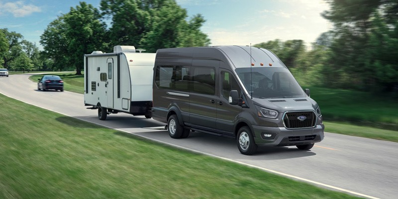 A Ford van towing a trailer on a road with green fields and trees
